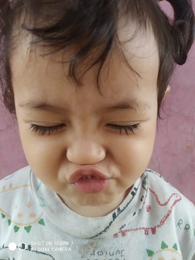 A young child's face is closely photographed. The child's eyes are closed and lips are pursed, creating a pouty expression. The child has dark hair and long eyelashes. They are wearing a white t-shirt with colorful cartoon designs. The background is a muted pinkish hue. The image includes text at the bottom indicating it was taken on a Redmi 7 AI Dual Camera.