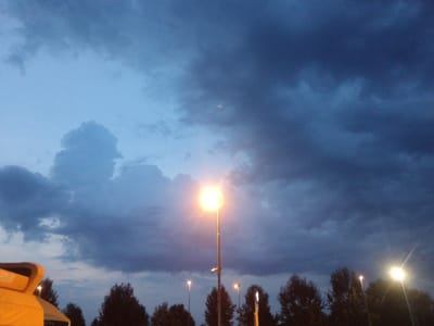This is a nighttime photograph of an outdoor scene, likely a parking lot or public space, with a dramatic sky dominated by dark, heavy clouds. The ambient light suggests dusk or early night. In the foreground, the left side of a bright yellow structure, possibly a tent or booth, is visible. Several streetlights are illuminated; one prominent streetlight in the center of the frame emits a bright, warm light. Other, less intense lights are visible in the background. Silhouetted trees line the horizon. The image does not contain any people or specific identifying landmarks to place it within the city of Alessandria, Italy.
