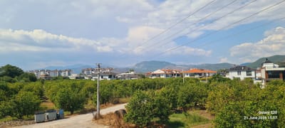 This image captures a landscape scene in Dalaman, Turkey, on June 21, 2026, at 15:57. The foreground features an unpaved dirt road with a row of green trees on either side, forming what appears to be an orchard or grove. Two metal trash bins are visible on the left side of the road. In the middle ground, more trees are present, interspersed with several buildings. These buildings are a mix of residential and commercial structures, with a prominent block of apartment buildings in the distance, some featuring satellite dishes on their roofs. Several houses with orange tiled roofs are also visible. A utility pole with power lines stretches across the upper portion of the image. In the background, rolling hills and mountains are present, with a few buildings scattered among them. The sky is partly cloudy, with large white cumulus clouds against a blue sky. The overall scene suggests a semi-urban or suburban environment within the Dalaman region, characterized by a blend of nature and development. The timestamp "Galaxy S20 21 June 2026 15:57" is overlaid on the bottom right corner.