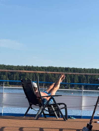 A person relaxes in a lounge chair on the deck of a boat, with their legs extended upward. The person is wearing blue swim trunks and a white cap, facing away from the camera. The boat is on a body of water, likely a lake or river, with a dense forest visible on the distant shore. The sky is clear and blue. The scene appears to be in Zavolzh'e, Russia, a city known for its proximity to large bodies of water and forests.
