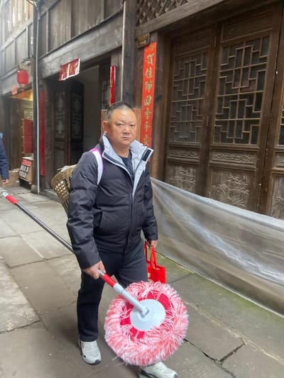 A man holds a mop with a red and white head. He wears a black jacket with a white collar, black pants, and white sneakers. He stands on a stone pathway next to a building with ornate wooden windows and a red banner. He carries a red plastic bag in his left hand, and a woven basket hangs off his left side. The image is rotated to the right.