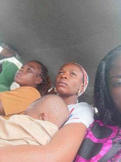 The image displays an interior scene, likely within a passenger vehicle, featuring five individuals. The vehicle's grey headliner is visible in the upper portion of the frame. In the foreground, on the right, a partial view of an individual with dark braided hair is present, wearing a pink and black patterned garment. This individual appears to be holding the camera.
Immediately to their left, a young woman is seated, wearing a white t-shirt and a light-colored headscarf with red and blue patterns. She has gold-colored hoop earrings and is looking upwards and to the left. Her right arm supports a sleeping infant. The infant, positioned centrally in the mid-ground, has a shaved head and is dressed in a light tan garment.
Behind and to the left of the infant, another young woman with dark braided hair is visible, wearing an orange-colored top and also looking upwards and to the left. In the far background, to the left, a partial view of a fifth individual is seen, wearing a green garment with an arm slightly raised. The specific location within Itobe, Nigeria, is not discernible from the visual information provided.