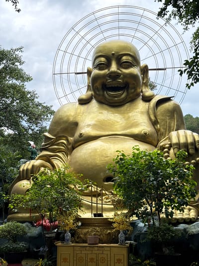 In Nam Ban, Vietnam, this image captures a colossal golden statue of the Laughing Buddha, or Hotei. The statue is situated outdoors, with lush green trees framing the massive figure. Behind the Buddha, a large circular metal structure is visible against a cloudy sky. In the foreground, decorative elements like incense sticks in a golden holder and potted plants with small yellow flowers add to the spiritual ambiance. To the left of the statue, a person dressed in green is seen from the side, holding a white fan. The statue's immense size and golden sheen dominate the scene, conveying a sense of grandeur and peace.