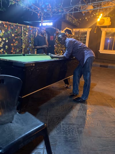 This image depicts a nighttime scene in the city of Igbessa, Nigeria, likely an outdoor recreational area or bar. Two adult males are present around a pool table.
In the foreground to the right, a dark-complexioned male, wearing a blue and white checkered long-sleeved shirt with rolled-up sleeves, blue jeans, and dark sandals, leans over the side of a black-framed pool table with a green felt surface. His right hand holds a dark smartphone above the table, and his index finger points towards a dark billiard ball and a white cue ball. His gaze is directed towards the table.
In the background to the left, a second dark-complexioned male, wearing a dark short-sleeved t-shirt, stands upright behind the pool table. He holds a dark smartphone to his right ear, appearing to be on a phone call, with his left hand holding another small dark object below the phone. He faces left, away from the camera.
The environment is illuminated by artificial lighting, including bright blue/white LED strip lights strung across an overhead trellis adorned with artificial leaves. A warmer yellow industrial-style light fixture is mounted on a dark building wall in the background right, featuring two decorative white window frames. A wall on the left is covered with artificial green and orange foliage. The ground is a dark, patterned surface, possibly stamped concrete or tiles. A dark, possibly plastic, chair is visible in the immediate lower left foreground. A few crumpled metallic objects lie on the floor under the pool table near the first man's feet.