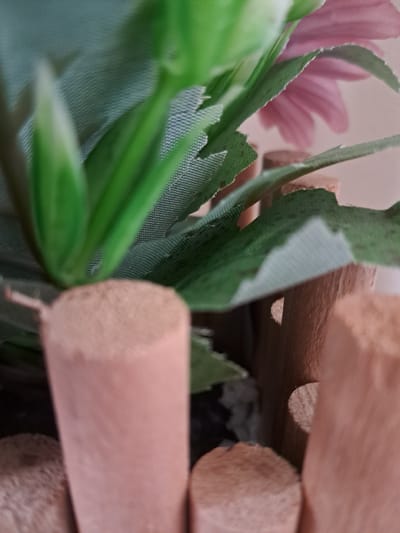 An extreme close-up shot captures an arrangement of artificial foliage, artificial flowers, and cylindrical wooden rods. In the foreground and center-left, bright artificial green leaves are prominent, characterized by their textured, mesh-like fabric and slightly frayed, jagged edges. Towards the upper-left, blurred pink petals of an artificial flower are visible in the soft-focus background, adding a delicate color contrast. On the right side and extending across the top, several light brown, roughly cut cylindrical wooden rods or dowels are stacked or arranged. Their ends show the natural wood grain and texture. The shallow depth of field emphasizes the foreground elements, blurring the background into soft hues. No specific activity is taking place, and the image depicts a static arrangement of decorative or craft materials. No text is visible. The scene is set in Gavar, Armenia, likely indoors with ambient lighting.