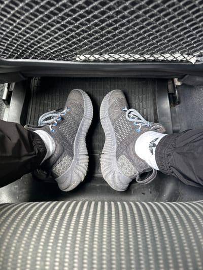 The image presents a downward, first-person view of two feet resting on a black rubber car floor mat. The feet are adorned with dark grey/black sneakers featuring light blue eyelets and speckled grey and white laces. The person wears white ankle socks, one of which prominently displays a black Adidas Trefoil logo. Black athletic-style pants, cinched at the ankles, cover the lower legs.

The scene is set inside a vehicle, likely a car or van, with the feet placed in the footwell. To the left, a black mesh storage net or pocket is visible, while to the right, a grey car seat fabric with a repetitive pattern of white dots fills the frame. The floor mat has a textured surface with raised patterns, typical for collecting debris. No specific activity is depicted beyond being present in the vehicle. The lighting suggests an interior environment. No emotions are discernible. The only visible text is the Adidas logo on the sock. The location is Zurzach, Switzerland.
