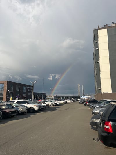 A rainbow arches across a dark, cloudy sky over a parking lot in front of a commercial building and a residential building. The parking lot is filled with parked cars. The commercial building has brick facade and multiple windows with signs. The residential building is tall and modern with a light-colored facade and balconies. The weather appears to be a mix of sun and rain, creating the rainbow.