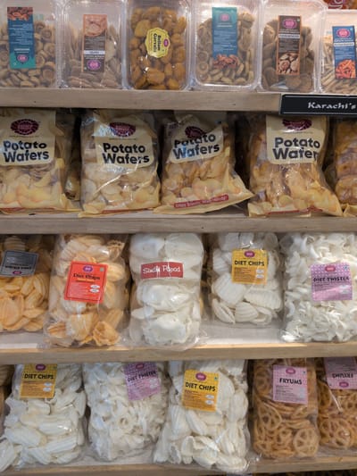 The image displays a retail store shelf unit featuring four tiers of packaged food products, predominantly from "Karachi Bakery," a brand established in 1953. Given the specific request for location, this scene is situated within a Karachi Bakery outlet or a retail section selling their products in Hyderabad, India.

The top shelf contains clear, rectangular plastic containers. From left to right, visible products include: "BAKARWADI," small, brown, spiraled snacks with a green vegetarian symbol; "PAPDI," flat, light brown, disc-shaped snacks; "Bellam Gavvalu," irregularly shaped, golden-yellow items; "PUDHINA MATURI PURI," dark brown, textured snacks; and partially visible "BLACK PEPPER MASALA STICKS," thin, elongated sticks. All these products carry the "Karachi Bakery" branding and typically a green vegetarian symbol.

The second shelf features a black sign with white lettering displaying "Karachi's" on the right side. Below this sign, three clear plastic bags of "Potato Wafers" are arranged side-by-side. Each bag contains light yellow, crinkle-cut potato chips and is labeled with "Karachi Bakery," "Quality & Goodness since 1953," and phrases like "Crispy, Crunchy, Totally Munchy!".

The third shelf holds various bagged snacks. From left to right: a partially obscured bag of wavy-cut chips; a bag of orange-yellow, crinkle-cut "Diet Chips" from Karachi Bakery, identified by a red label; a bag of white, irregular, shell-shaped items labeled "Sindhi Papad" directly on the bag; a bag of white, crescent-shaped "Diet Chips" from Karachi Bakery, with a yellow label; and a bag of white, twisted, spiral-shaped "DIET TWISTER" from Karachi Bakery, identified by a purple label. All Karachi Bakery branded products on this shelf feature a green vegetarian symbol.

The fourth and bottom-most visible shelf continues the display of bagged products. From left to right: a bag of white, irregular, shell-shaped "Diet Chips" from Karachi Bakery, with a yellow label and green vegetarian symbol (identical to one on the third shelf); a bag of brown, circular, lattice-patterned snacks labeled "FRYUMS" from Karachi Bakery, with a purple label and green vegetarian symbol; and another identical bag of "FRYUMS" to its right.

The shelves are constructed of light-colored wood. No individuals are present in the frame. The lighting is uniform, suggesting an indoor commercial environment.