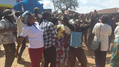 A jubilant crowd gathers outdoors under a bright sky. A woman with a blue headwrap and white shirt leans back with a smile, while a man beside her in a checkered shirt looks towards the center. Another man, with arms raised, wears a blue and black shirt. A building roof is visible in the background. The scene conveys excitement and celebration.