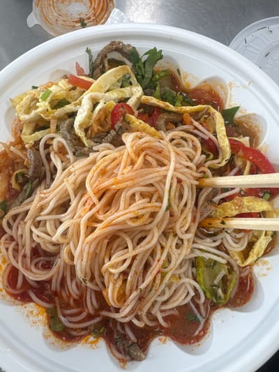 A close-up, overhead shot shows a white, circular bowl filled with a noodle dish. The noodles are thin and white, coated in a reddish-orange sauce with visible flecks of spice. Interspersed with the noodles are slices of fried egg, thin strips of cooked meat, chopped tomatoes, green herbs, and possibly some bell pepper. Chopsticks are holding a portion of the noodles. The dish appears to be served in a disposable takeout container. In the background, to the upper left, is another small disposable container with a similar reddish sauce and lid. The dish is placed on a metal surface, likely a table. This dish is a representative example of food commonly available at food stalls or restaurants in Hwaseong-si, South Korea.