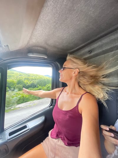 A woman with blonde hair and sunglasses is leaning out of a vehicle window, her arm extended towards the outside. Her hair is blowing in the wind. She is wearing a maroon tank top and light-colored shorts. The interior of the vehicle, including the ceiling and a door panel, is visible. Outside the window, a lush green, tree-covered hillside under a bright sky is seen, suggesting a rural or mountainous location. The specific location in Ol'ginka, Russia cannot be determined from the visual information alone.