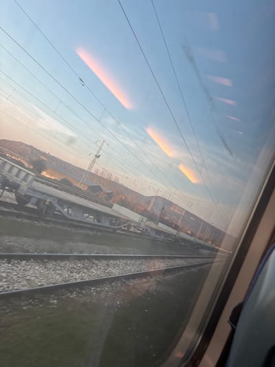 The image depicts a railway scene in Brusen, Bulgaria, viewed from inside a moving train, looking out a window. The perspective is slightly angled downwards and to the left. The upper portion of the frame shows a clear, light blue sky with elongated, horizontal reflections of warm orange and pink light, likely from the sun, appearing as bright streaks across the windowpane. Multiple overhead electrical lines (catenary wires) traverse the sky, supported by at least one visible utility pole in the mid-ground.
Below the horizon, an industrial or rural landscape is visible. On an adjacent track parallel to the viewer's train, a long freight train or series of disconnected freight wagons are present. These include flatcar-type wagons and open-top wagons, some appearing to contain bulk material. In the background, a gently sloping hill or mountain range is illuminated by warm, low-angle sunlight, creating an orange hue on its face. Scattered structures, possibly industrial buildings with dark roofs, are situated at the base of this distant landform. The immediate foreground, outside the window, consists of multiple parallel train tracks laid on gravel ballast, bordered by patches of green ground cover. No individuals are discernible.