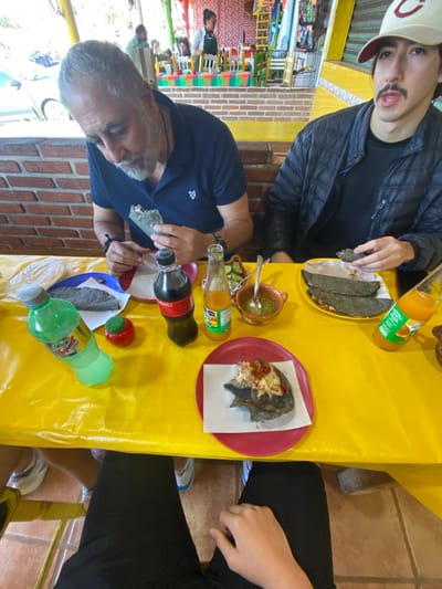 Two men are seated at a yellow table laden with Mexican food and beverages. Plates hold what appear to be quesadillas. Various bottles of soda and juice are scattered across the table, along with a salsa dish. One man wears a navy shirt, while the other sports a cap and a black jacket. The setting suggests a casual dining experience.