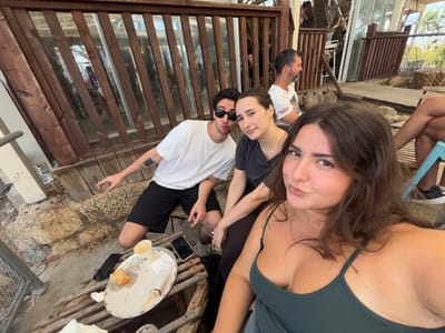 An outdoor scene in Kfar Vitkin, Israel, depicts a group of individuals on a patio. In the immediate foreground, a woman with dark hair, wearing a dark green tank top, looks directly at the camera with a slight facial expression. Behind and to her left, a man with dark hair, sunglasses, a white t-shirt, and black shorts is seated, extending his right arm with an open hand gesture. A dark square tattoo is visible on his left forearm. Seated next to him, a woman with dark hair, wearing a dark grey t-shirt, looks towards the camera.
In the mid-ground, a man with a grey beard and a white t-shirt is visible, seated and facing to the right. On the far right, the lower legs and feet of another individual are partially visible, seated on a light blue stool.
In the lower left foreground, a small, round white table holds an oval tray containing an empty paper cup, remnants of a pastry, and a dark-colored cell phone. The ground consists of rough stone paving and wooden deck planks. The background features wooden slatted barriers, glass window panes reflecting indistinct figures and the exterior, and glimpses of natural light and sparse vegetation.