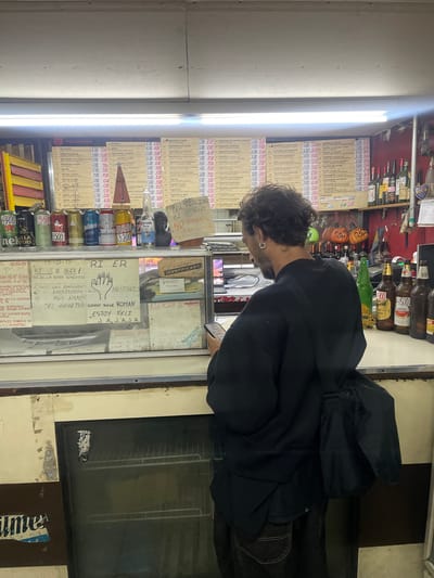 A man stands in front of a storefront in Vicente López, Argentina. He is looking at a handwritten menu and a glass display case filled with canned goods and price lists. The sign on the case has handwritten notes that include "RI ER O BUCA 1," "Falto Gas PIFICENTA", and "ESTOY FELI JA JAJA". The menu board on the left lists various pizza toppings and prices. The store also sells beer and other drinks. The storefront has a window that is lit by a fluorescent light. A refrigerator with a glass door is visible on the right. A partially visible street sign says "aume". The location is most likely in the province of Buenos Aires, Argentina.