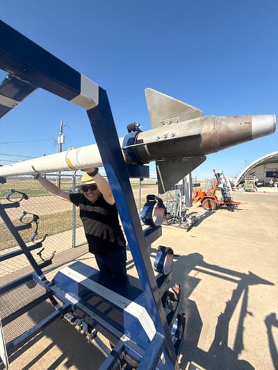 A missile stands on a blue and white gantry. A man in a black shirt, jeans, and a yellow hard hat holds onto the gantry. He is smiling and wearing sunglasses. The missile is silver and grey. The ground is concrete. A hangar and other equipment sit in the background. The sky is blue and cloudless.