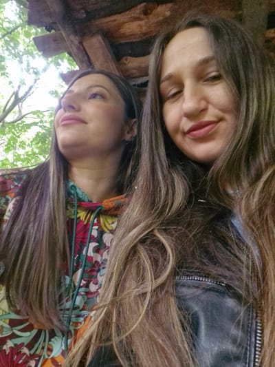 Two women are captured in a selfie-style shot. The woman at the top is wearing a black leather jacket, and the one below is wearing a multi-colored floral patterned shirt. The location appears to be outdoors, possibly a park or wooded area in Krichim, Bulgaria, as evidenced by the presence of greenery and wooden structures in the background.
