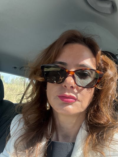 A woman with brown hair and sunglasses is photographed from a low angle. She is wearing a white jacket over a black top. The photograph was taken inside a vehicle, with the interior visible on the left side. Outside, a blurred landscape is visible through the car window. While the location is identified as Pombal, Portugal, the image doesn't provide specific landmarks or identifiers that would confirm her exact location within the city.