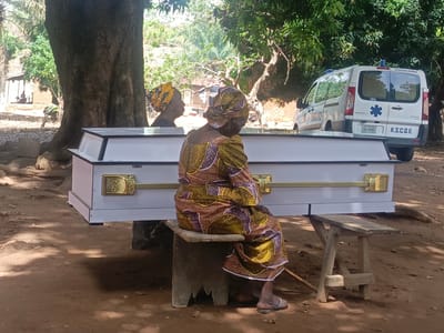 The image depicts a solemn scene in Ninzo South, Nigeria. In the foreground, an elderly woman dressed in a patterned brown and gold outfit sits on a low concrete stool. Her back is to the viewer, and she rests her arms on a white casket with gold handles. Beside her is another woman wearing a yellow headwrap whose face is partially visible. Behind the casket is a parked white ambulance with the words "AMBULANCE" and "K.S.C.O.E." visible. The scene is shaded by large trees. The ground is covered in reddish-brown earth. In the distance, a small structure is visible.
