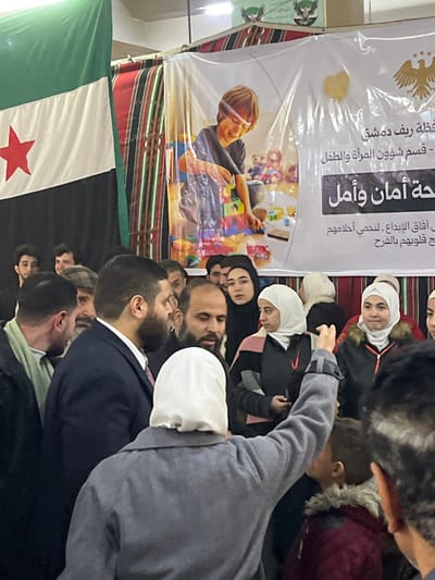 An indoor scene in Kafr Batna, Syria, depicts a dense gathering of approximately 20-30 individuals, primarily adults. In the immediate foreground, an individual wearing a grey coat and a white head covering is seen from behind, with their right arm raised vertically above the crowd. To their right, the back of a child's head, wearing a brown hood with fur trim, is visible.
The mid-ground features a tightly packed group of men and women. Several men with beards are present, one in a dark suit jacket, while numerous women wear hijabs; some are observing attentively, others are smiling or engaging with unseen activity.
The background displays a large flag on the left, characterized by green, white, and black horizontal stripes with a red five-pointed star centered on the white stripe (the Syrian opposition flag). Adjacent to this, a large white banner features a graphic of a smiling child playing with colorful building blocks and toys. Arabic text on the banner identifies it as relating to the "Damascus Countryside Governorate," a "Women and Child Affairs Department," and phrases translating to "Safety and Hope." Two golden eagle emblems are visible on the upper right of this banner. Behind the banner, striped fabric in red, black, and white is discernible, suggesting decorative drapery or a tent structure. Overhead lighting fixtures are partially visible at the top of the frame.