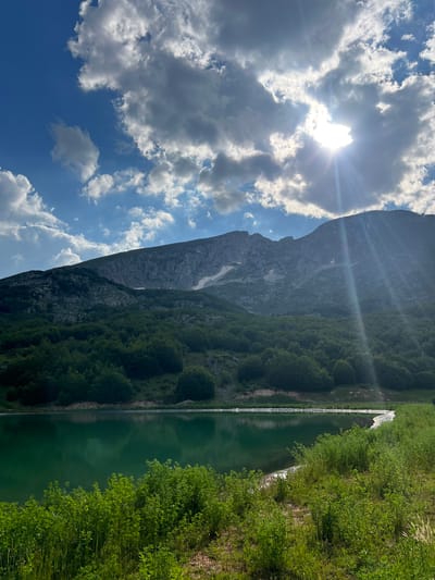 This is a vertical, eye-level, outdoor shot of a scenic mountain landscape with a lake. The sun is shining brightly, with its rays visible through the clouds and casting lens flares across the image. The sky is blue with scattered white and gray clouds. A large, dark mountain dominates the background, with patches of snow visible on its slopes and a dense green forest covering its lower sections. To the right of the mountain, a calm, green lake stretches into the distance, bordered by lush green vegetation and trees. The reflection of the sky and the mountain is visible on the water's surface. The time of day appears to be daytime, likely late morning or early afternoon, given the bright sunlight. The weather is clear and sunny. The location is Motički gaj, Montenegro, a natural setting characterized by mountains, forests, and a lake. There are no people or discernible objects apart from the natural elements. The overall mood conveyed is one of tranquility and natural beauty.