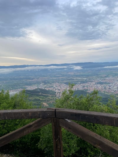 An elevated, wide-angle shot captures a scenic overview of a city nestled in a valley, viewed from a wooden railing. The sky above is overcast with diffused light, indicating either early morning or late afternoon. In the foreground, a weathered wooden railing made of thick, dark timbers forms a protective barrier. Some of the timbers bear graffiti and carvings. Lush green trees with light-colored blossoms are visible behind and around the railing.
The mid-ground reveals a sprawling urban landscape with densely packed buildings featuring red tile roofs. The city extends into the valley, interspersed with patches of green. Rolling hills and mountains form the backdrop, with a layer of low-lying clouds or fog partially obscuring the distant mountain range. The overall impression is one of a peaceful, vast panorama overlooking a settled area. The specific location is not identifiable from the visual information alone; it is a generic representation of a city in a valley surrounded by nature.