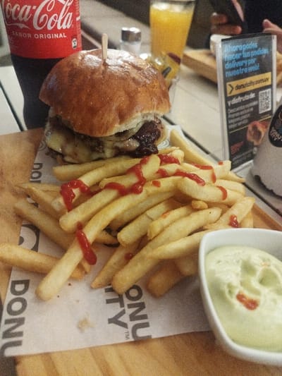 A burger with a toothpick, covered in cheese, sits atop a pile of french fries drizzled with ketchup on a branded paper. A small bowl of greenish dip accompanies the fries. A Coca-Cola bottle stands next to the burger, and an orange juice glass is visible in the background. A sign promoting donutcity.comsuy is also present, along with a partially visible salt shaker. The scene appears to be a meal at a restaurant.