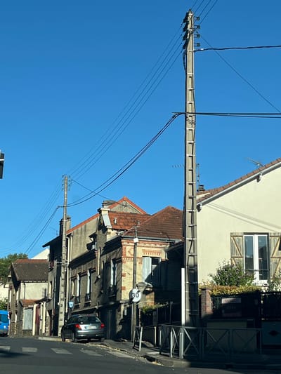 The image shows a street scene in L'Haÿ-les-Roses, France. The view is from a street level, looking down a road lined with buildings and utility poles. The sky is clear and blue.
In the foreground, a dark gray car is parked on the right side of the road, partially obscuring the sidewalk. A speed limit sign indicating "30" is visible on the right.
The buildings are a mix of architectural styles, with some appearing older and others more modern. The building on the far right has a cream-colored facade, wooden shutters on a window, and a "VENDU" (sold) sign in front of a planter. Adjacent to it are older buildings with brick and stone facades, and tiled roofs. Utility poles with extensive wiring are prominent throughout the scene, a common feature in many urban and suburban environments.
On the left side of the road, a blue van is partially visible. The road surface appears to be asphalt with painted lane markings and a pedestrian crossing. The overall impression is a typical residential or mixed-use street in a French town.