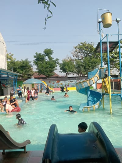 The image depicts a sunny day at a public swimming pool with a water slide and splash bucket attraction in Kemamang, Indonesia. Several children and adults are enjoying the water. In the foreground, a child is visible near a dark blue slide. In the mid-ground, a group of children are playing in the shallow end of the pool, with some standing and others sitting on the edge. A blue and yellow water slide curves into the pool, and a large yellow splash bucket is positioned above the slide. A wall with colorful underwater-themed murals lines the back of the pool area. Trees and buildings are visible in the background under a clear blue sky.