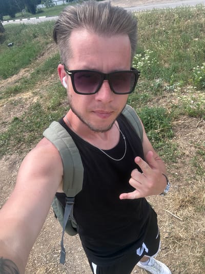 A young man with light brown hair, sunglasses, and a black tank top is taking a selfie outdoors. He is wearing white earbuds, a silver chain necklace, and a light green backpack. He is making a rock-on hand gesture with his right hand. The background consists of dry grass, dirt, and some greenery, suggesting a rural or park-like setting. The location is identified as Aeroflotsky, Ukraine.