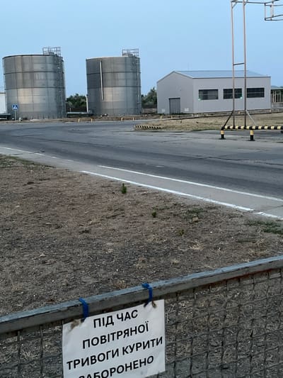 The image shows an industrial area with two large cylindrical storage tanks on the left, a grey warehouse-like building in the background, and a paved road in the foreground. The tanks are made of metal and have ladders on their sides. The building has a silver roof and several windows. A yellow and black striped barrier is visible on the right side of the road. In the foreground, there is a metal fence with a sign attached to it. The sign contains text in Ukrainian which translates to "During the air alarm, smoking is prohibited." The scene appears to be set in the city of Mykolaivske, Ukraine, given the context provided. The sky is overcast and the overall lighting suggests it is either early morning or late evening.
