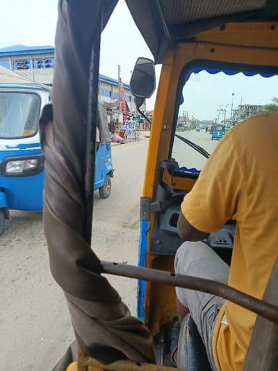 The image captures a daytime street scene from the interior perspective of a yellow commercial tricycle, common in Warri, Nigeria. The driver, wearing a mustard yellow t-shirt and light grey shorts, occupies the right side of the frame, with his right arm bent and hands likely on the steering mechanism. The road ahead is unpaved, consisting of reddish-brown dirt or gravel, extending into the distance where several blue tricycles are visible.
To the immediate left, a white and blue tricycle is stationary or moving slowly. Further along the left side of the road, a blue tricycle is parked near a series of roadside stalls or small shops operating under awnings. A white sign with "POS" in blue lettering is prominently displayed, indicating a point-of-sale service. Behind these shops, a tall red and white striped utility pole or antenna stands next to a multi-story building featuring a blue corrugated metal roof and numerous small, dark-framed windows. The sky overhead is a hazy, light blue.