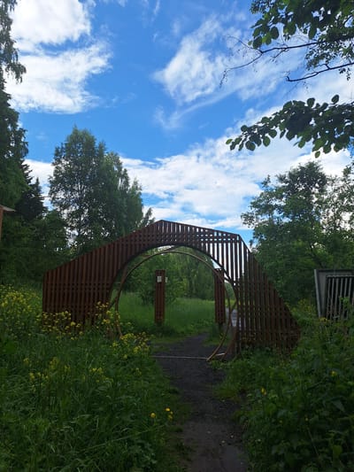 The image depicts an outdoor scene in the city of Sim, Russia. In the foreground, a gravel path leads towards a large, circular wooden archway. The archway is constructed of vertical wooden slats and appears to be part of an entrance or gateway. Surrounding the path and archway is lush green vegetation, with tall grasses and scattered yellow wildflowers.
Beyond the archway, the path continues into a wooded area with numerous trees and foliage. The sky above is a bright blue, dotted with fluffy white clouds. To the right of the archway, a metallic structure with vertical bars is partially visible, suggesting it might be part of a fence or enclosure. The overall impression is of a natural, park-like setting, possibly an entrance to a trail or a recreational area.