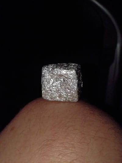 A cube wrapped in aluminum foil is held against a fair-skinned human arm. The background is dark. There is no location information available.