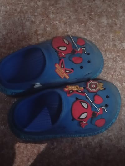 Two blue Spider-Man themed children's slides are displayed on a textured, mottled brown surface, possibly a floor or ground. The slides feature prominent red and white Spider-Man masks and branding, with the words "SPIDER-MAN MARVEL" visible on one slide. The slides are positioned next to each other, with the top slide slightly offset from the bottom one. The lighting suggests an indoor environment. There are no people present in the image. The scene is a still life presentation of the footwear. The location is specified as Zhany-Zher, Kyrgyzstan.