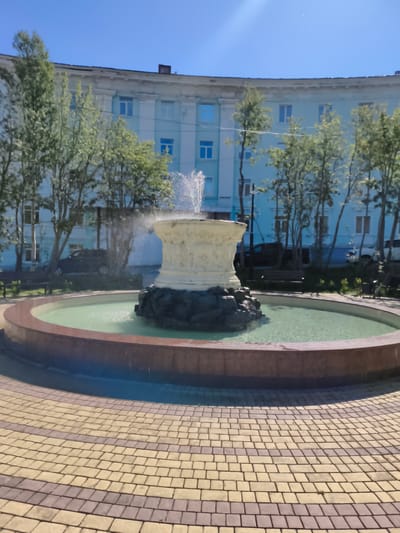 A circular fountain with a light-colored ornate basin and a dark rocky base is at the center of a plaza paved with tan and purple interlocking bricks. The fountain is active, with water spraying from its top and misting around its base, creating a green hue in the shallow pool. Behind the fountain, a light blue building with white trim and numerous windows curves along the plaza's edge. Several trees with sparse green foliage are planted in front of the building, some obscuring the lower floors. Benches are visible under the trees and behind the fountain. The sky is a clear, bright blue, suggesting a sunny day. The scene appears to be in an urban setting, possibly a public square or park. This location is confirmed to be the Fountain Square in Kirovsk, Russia.