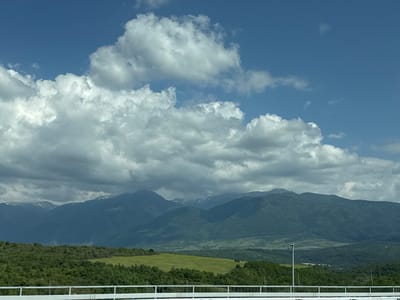 This landscape image captures a panoramic view of layered mountains under a dynamic sky. The scene is dominated by a succession of dark green, forested mountain ranges, stretching across the mid-ground and receding into the distance. Several higher peaks exhibit small patches of snow or lighter rock, suggesting altitude.
Above the mountains, the sky displays a blend of clear blue areas and prominent, fluffy cumulus clouds. These clouds vary in size and density, ranging from bright white to soft grey, and are scattered across the upper and mid-sections of the sky.
In the mid-foreground, undulating green hills and fields create pockets of bright greenery amidst the darker forested slopes. Dense foliage and trees also fill the immediate foreground.
At the bottom of the frame, a white metal barrier or railing, characteristic of a bridge or roadside, runs horizontally. A tall, slender light pole is visible on the lower right, indicating human infrastructure.
The image depicts a bright daytime scene, likely under partly cloudy conditions. No people or visible text are present. The setting is a scenic, mountainous area, consistent with the location context of Balanovo, Bulgaria.