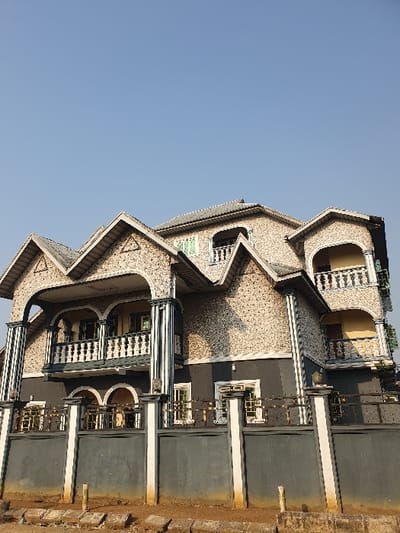 An angled perspective of a multi-story building against a clear blue sky. The building's facade features detailed stone work, arched windows, and decorative balconies. A portion of the rooftop and its railings are visible. The building's right side reveals a darker section with rectangular structures and metal railings. The ground appears as a blurred, orange-toned surface.