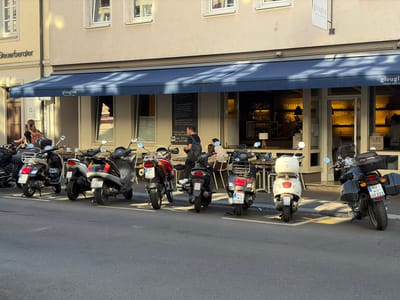 A row of parked scooters and motorcycles lines a street in front of a building with a blue awning in Würzburg, Germany. The building appears to be a restaurant or cafe with outdoor seating. A sign above the awning reads "glouglou RESTAURANT | WEINBAR | BISTROT". Several people are seated at tables under the awning, and some are walking in front of the building. The street is paved, and there are yellow parking lines painted on the ground. The sun is shining, casting shadows on the street and building. The scooters and motorcycles are of various makes and models, and they are all parked facing the street. Some of the license plates are visible, with some displaying "WU" which is the code for Würzburg.