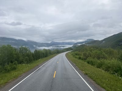The image depicts a winding asphalt road with a white line marking its edges and yellow dashed lines indicating lanes. The road is surrounded by lush green grass and dense foliage on either side, leading towards a backdrop of majestic, mist-shrouded mountains. The sky is overcast with gray clouds, contributing to a moody, serene atmosphere. The scene is consistent with the landscape found in Sørfjordmoen, Norway, characterized by its dramatic fjords, mountains, and verdant vegetation. There are no people or vehicles visible in the image, focusing solely on the natural environment and the man-made road traversing it. The overall impression is one of a peaceful, remote journey through a picturesque natural setting.