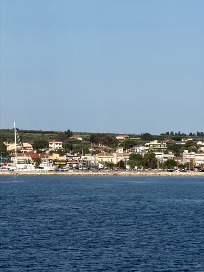 The image depicts a coastal town in the Municipal Unit of Kastro - Kyllini, Greece, viewed from the water. In the foreground, the deep blue sea stretches across the frame, with visible ripples on its surface. Along the shoreline, a quay is lined with a variety of boats, including a prominent white sailboat with its mast extending high into the sky. Behind the quay, the town unfolds with a cluster of buildings, characterized by red-tiled roofs and a mix of architectural styles. Trees and vegetation are interspersed among the structures, suggesting a Mediterranean landscape. The background is dominated by a clear, pale blue sky. Along the waterfront, there is a street with parked cars, indicating a functioning port or harbor area.