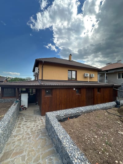The image depicts a residential property in Gorna Grashtitsa, Bulgaria, under apparent landscaping or construction. A two-story house with an orange-ochre facade and a brown tiled roof occupies the center-left. Two external air conditioning units are mounted on its upper wall, alongside a dark-framed window. A brown chimney with a light shaft extends from the roof. Attached to the main structure is a single-story extension or separate building with walls clad in vertical dark wooden planks, covered by a matching brown tiled roof. This wooden structure features multiple small, dark-framed windows and a dark-framed door. Under a covered section of this extension, a white refrigerator, a red chainsaw, and other tools are visible on a concrete surface.

To the right, a partially visible neighboring house features a grey facade, a brown tiled roof, a balcony with a metal railing, and an external staircase. Two air conditioning units are also mounted on its exterior.

The foreground consists of a meticulously laid pathway made of irregular, flat flagstones set in mortar. This pathway is bordered on both sides by low retaining walls constructed from gabion baskets filled with light grey crushed stone. To the right of the pathway and the wooden structure, an open area of bare, disturbed soil with scattered small rocks and sparse green vegetation is present, also partially enclosed by gabion walls. A black irrigation hose and orange marking pegs are visible in this soil area.

The sky is a prominent element, transitioning from clear, bright blue on the left to a dense accumulation of white and grey clouds on the right, with patches of blue visible through breaks in the cloud cover. No individuals or animals are present in the scene.