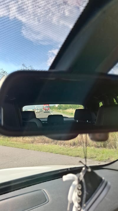 The image shows a view through a car's rearview mirror, looking towards the rear windshield and the road beyond. The scene is set on a highway, with a red truck and several other vehicles visible in the distance. The road is paved and flanked by grassy verges and trees. The sky is blue with scattered clouds. In the foreground, parts of the car's interior are visible, including the dashboard, the rearview mirror itself, and a novelty car air freshener shaped like a miniature sneaker hanging from the mirror. The location is identified as Kalugerovo, Bulgaria.