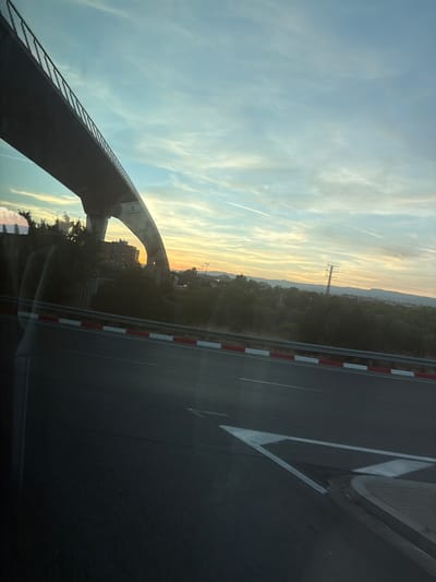 The image captures a road adjacent to a bridge structure with a natural landscape and sky in the background. The time of day appears to be sunset or sunrise, given the warm, golden hues in the sky and the soft lighting. The sky is a blend of light blue and yellow, with wispy clouds.
In the foreground, a dark asphalt road is visible, marked with white lines and bordered by a red and white striped barrier. The left side of the image is dominated by the underside of a large, modern bridge structure, which is dark and appears to be made of concrete or steel. The bridge arches over a natural area that includes trees and some buildings. A tall electrical pylon can be seen in the middle distance, silhouetted against the colorful sky.
The landscape beyond the immediate road and bridge features rolling hills or mountains in the far distance. There are hints of urban development with buildings visible amongst the trees. The overall mood is serene and perhaps a little melancholic, consistent with a twilight setting.
No people are visible in the image. The visible text appears to be a white label or sign on the underside of the bridge, but the text itself is illegible. The setting, with the bridge, road, and distant landscape, suggests an area on the outskirts of a town or city, likely in a developed region given the infrastructure. The weather appears to be clear, with the exception of the clouds. The location context provided is Vila-seca, Spain, which is consistent with the type of infrastructure and landscape depicted.