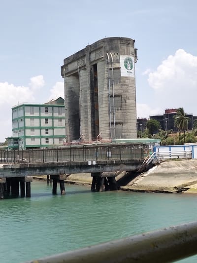 The image depicts an industrial waterfront scene under a clear sky with scattered white clouds, located in Mkomani ward, Kenya. In the midground, a large, weathered concrete industrial facility dominates the right side. This structure features multiple vertical sections, a prominent curved cylindrical component on its right, and displays a "Bamburi" logo with a green tree emblem. Ladders and maintenance platforms are visible on its exterior. To the left of this, another multi-story building with light green walls and numerous white-framed windows is situated.
Separating these land-based structures from a body of turquoise water in the foreground is a concrete pier or bridge, supported by multiple robust concrete pillars submerged in the water. This structure features a dark metal fence or railing along its top surface. Adjacent to the large concrete facility, above the pier, a platform with reddish-orange safety railings is visible.
On the far right in the background, a cluster of large industrial storage tanks, some dark and some with red tops, are visible on an elevated landmass, alongside a solitary palm tree and other vegetation. A white and blue structure is also visible on the land near the water's edge, adjacent to a concrete embankment. The immediate foreground shows a blurry, dark metallic rail extending horizontally across the bottom of the frame, indicating the vantage point is from a watercraft or elevated platform. No individuals or active interactions are visible in the scene.