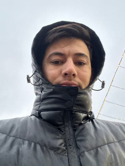 The image depicts a male individual, appearing to be in his twenties or thirties, centered in a close-up, upward-angled shot, likely a selfie, against an overcast sky in Yangiiul, Uzbekistan. He has short, dark hair and a neutral facial expression, with visible eyes, nose, and mouth, and slight stubble. He is wearing a dark, possibly black, puffy hooded jacket with the hood up. The jacket's high collar is zipped and fastened with two visible black snaps or buttons, covering his neck and lower chin. Drawstrings with black toggles are visible on either side of the hood. The jacket's surface, particularly on the shoulders and collar, shows evidence of moisture or droplets. The background consists of a uniformly light grey, cloudy sky. To the upper right, a vertical structure adorned with numerous small, warm-toned lights is partially visible, indicating an outdoor decorative light installation. The overall environment appears damp or wet, consistent with cloudy weather.