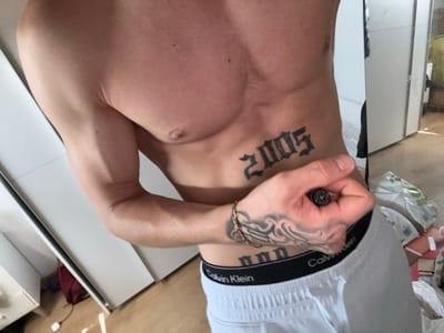A medium shot captures a person's torso and left arm, prominently displaying tattoos and Calvin Klein underwear. The tattoo on the abdomen reads "2005" in a gothic font, while another on the forearm appears to be tribal-inspired waves. The individual is wearing white boxer briefs with the Calvin Klein logo. Their left hand is visible, holding what appears to be a small, cylindrical object. The background suggests an indoor setting with white doors and wooden flooring, possibly a bedroom or changing room. The lighting suggests a bright, possibly sunlit, environment. The overall scene is presented in a forensic, matter-of-fact style, devoid of any emotional or subjective interpretation. No specific location in Villa Del Prado, Spain is discernible from the visual information provided.