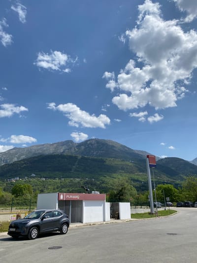 The image displays a sunny daytime scene in a mountainous region, likely Kavač, Montenegro. In the foreground, a dark gray SUV, resembling a Toyota C-HR, is parked on an asphalt lot. Its license plate, "KVB B...902", is partially visible, indicating a Montenegrin registration. To the right of the car is a modern, white building with a red and gray facade. On the building, a sign displays "PUNJENJE" with an icon of a bottle/cylinder, and "AUTOGAS" below it, suggesting it's an LPG or gas filling station. Near the parked car, a yellow warning sign is visible with symbols for caution and fire hazard, and the Cyrillic text "A ЗАБРАНЬЕНО" (Forbidden).

Further back, a tall signpost features a red section with the word "PETROL" and a blue section above it. The background is dominated by large, lush green mountains, characteristic of the Montenegrin landscape. The sky is bright blue with scattered white cumulus clouds. The lighting suggests it is late morning or early afternoon. The scene appears calm and clear, with no people visible. The asphalt area extends into a road or parking lot with green verges and other parked vehicles partially visible in the distance among the trees.