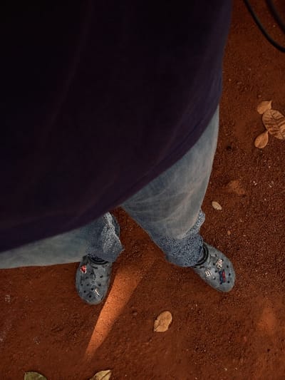 A high-angle view captures the lower body of an individual standing on reddish-brown earth in Oturkpo, Nigeria. The person wears a dark blue top, light blue denim jeans, and dark grey Crocs. The left Croc features a red circular charm and a silver-colored charm, while the right Croc displays a blue and red emblem along with a silver capital "D" charm. The ground, composed of fine reddish-brown soil, is sparsely covered with scattered dried brown leaves. A distinct diagonal band of sunlight illuminates the ground immediately surrounding the feet and extending forward.