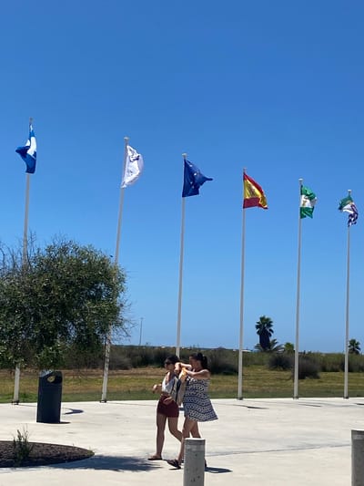The image depicts an outdoor scene under a clear blue sky, likely in or near Conil de la Frontera, Spain, given the flags present. In the foreground, two individuals, appearing to be young women, are walking on a paved surface. One is wearing a white top and maroon shorts, while the other is in a patterned dress. Behind them, several flagpoles are erected, each flying a different flag. The flags visible include what appears to be the European Union flag, the Spanish flag, and the flag of Andalusia. There is also a white flag with some text and a blue and white flag. To the left, a dark green trash receptacle is situated near a leafy tree. The background consists of dry vegetation and a glimpse of palm trees, suggesting a warm, coastal climate. The overall impression is one of a public space, possibly near a coastal area or a civic center.